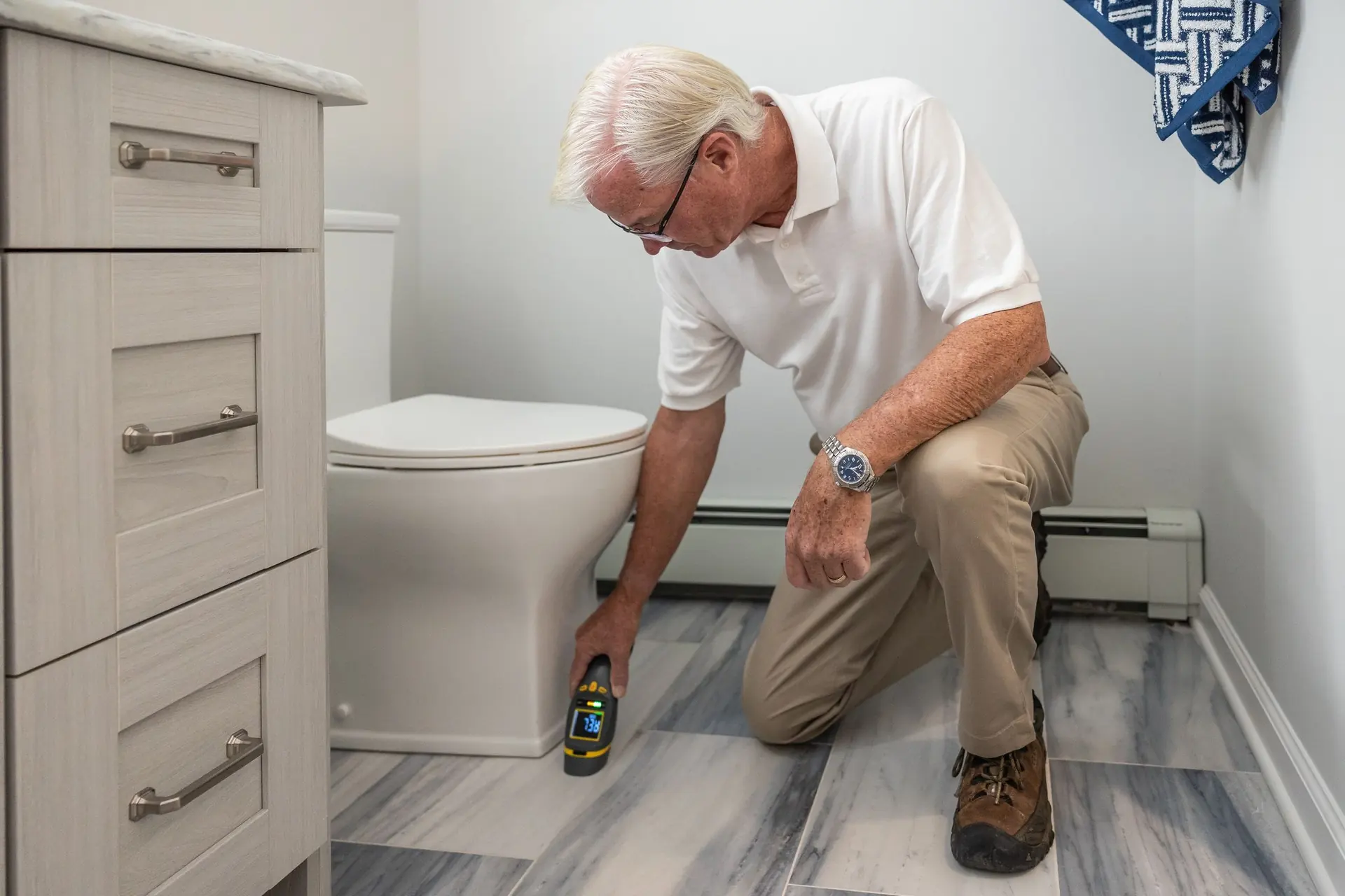 A grey-haired technician kneels on a bathroom floor, holding a moisture meter against the tiles beside the toilet.