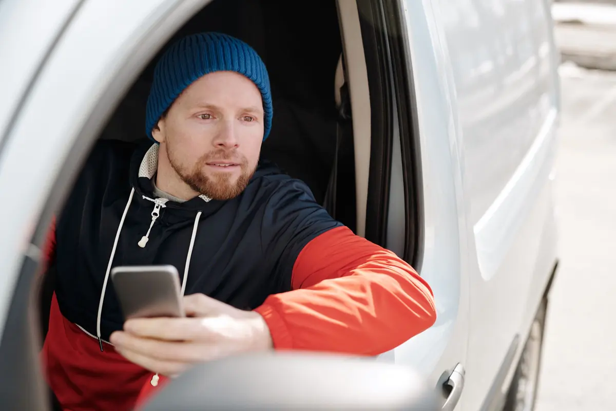 A man in a blue beanie sits in the driver's seat of a white van, phone in hand, glancing out of the window.